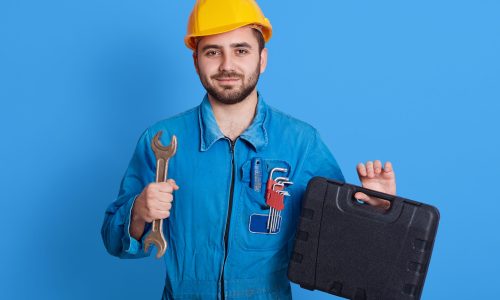 Young handsome repairman in blue overall and yellow helmet holding toolbox and wrench, bearded plumber standing isolated over color wall, man working, holds toolbox with instrument.