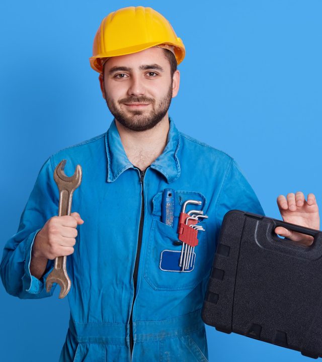 Young handsome repairman in blue overall and yellow helmet holding toolbox and wrench, bearded plumber standing isolated over color wall, man working, holds toolbox with instrument.