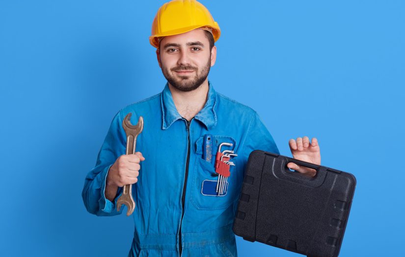 Young handsome repairman in blue overall and yellow helmet holding toolbox and wrench, bearded plumber standing isolated over color wall, man working, holds toolbox with instrument.